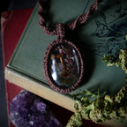 A handmade macrame pendant necklace with a clear oval case containing dried mushrooms and purple flowers, resting on an old green book next to a purple crystal cluster.